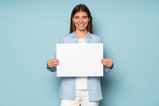 Lovely female executive manager holding white blank banner with copy space for your content or text, feeling successful and proud with her job isolated over blue studio background