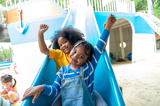 Happy Little African child boy and girl friends sliding and playing at outdoor playground in the park on summer vacation. Kindergarten children kid enjoy and fun outdoor activity learning and exercising at park.