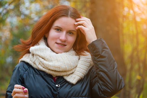 portrait of a red-haired smiling girl in a jacket and scarf straightening her hair. against the background of autumn nature and the bright sun, the concept of human emotion.