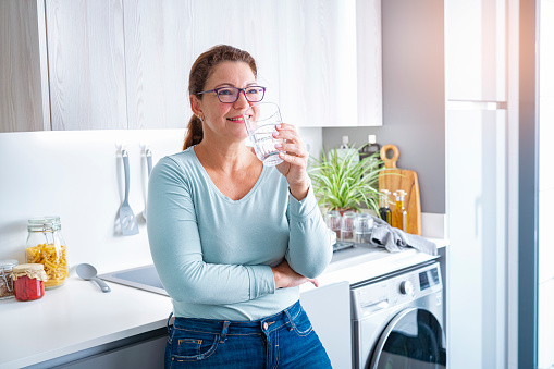 Side view of a mature woman holding a glass of fresh drinking water in modern kitchen. High resolution 42Mp indoors digital capture taken with SONY A7rII and Zeiss Batis 40mm F2.0 CF lens