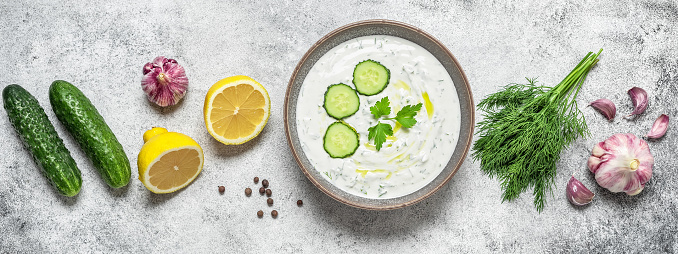 Tzatziki sauce in a bowl with ingredients. Gray rustic background. Traditional Greek sauce made from yogurt and vegetables. Top view, flat lay. Banner