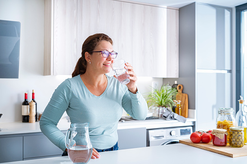 Smiling mature woman drinking water in modern kitchen. High resolution 42Mp indoors digital capture taken with SONY A7rII and Zeiss Batis 40mm F2.0 CF lens