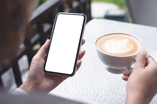 Cell phone mockup image blank white screen. Woman hand holding texting using mobile phone and cup of coffee at home.