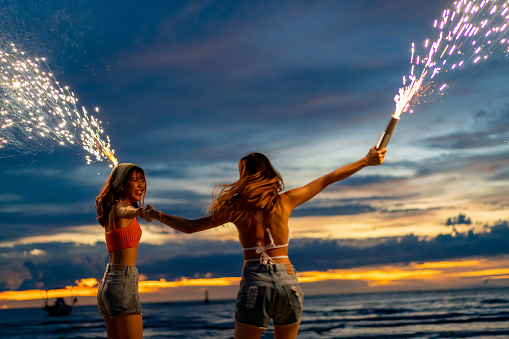 Young beautiful Asian woman having fun dancing and playing sparklers together on tropical island beach in summer night. Happy female friends enjoy outdoor lifestyle nightlife on holiday travel vacation trip