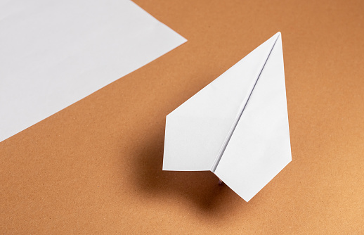 White origami plane and paper sheet on brown background with shadow. Handmade flying construction. High quality photo
