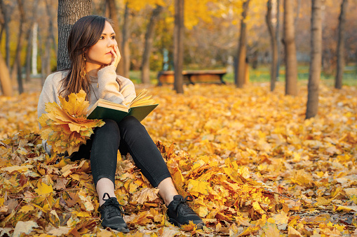 A young woman sits with a book in her hands on autumn foliage near a tree and dreams about something.