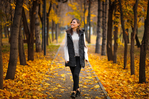 A young woman walks in an autumn park along an alley with fallen autumn leaves.