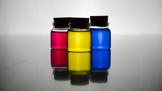 Three glass jars with cork stoppers with the three primary colors, magenta, blue and yellow in a backlight that are reflected on a mirror surface