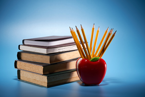 apple shaped pencil holder with pencils with stack of books against blue background