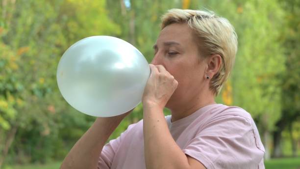 fille femme aux cheveux blonds courts gonfle un ballon sur fond de nature verte en été - inflating balloon blowing air photos et images de collection