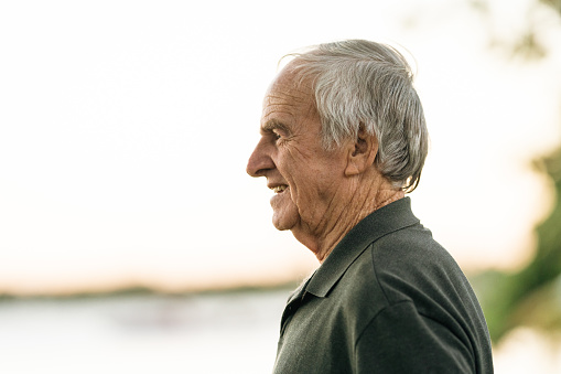 Portrait of happy retired senior man standing at home near window. Satisfied old man in casual clothes looking at camera and smiling while standing near the window. Positive and confident elderly enjoy his retirement at care facility.