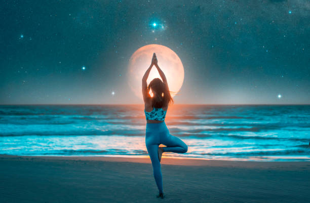 silhouette of a woman doing yoga on the shore of the beach with the moon in the background stock photo