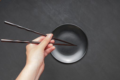 Pair of lacquered chopsticks in hand over an empty matte black plate on dark background.