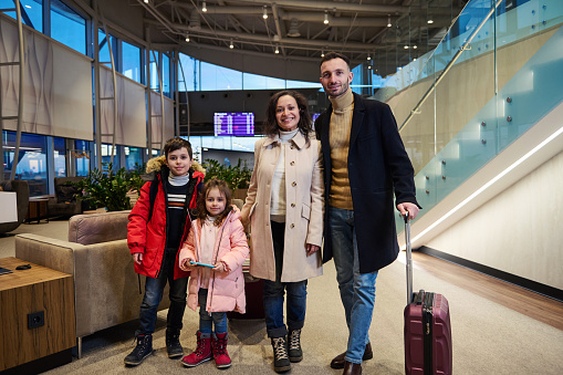 Cheerful family with suitcases, against flight information board with timetable in international airport departures terminal. Air travel concept, family relationships, children and parents together