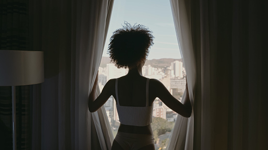 Beautiful Latin woman waking up in the morning, the sun shines on her from the big window. Happy young woman greets the new day with warm sunlight and city scenery in the window.
