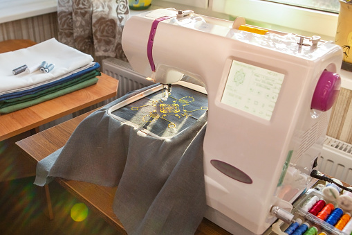 Home sewing workshop, workplace opposite the window, with a modern embroidery machine that creates a yellow pattern on a gray linen fabric. Next to the thread and fabric.