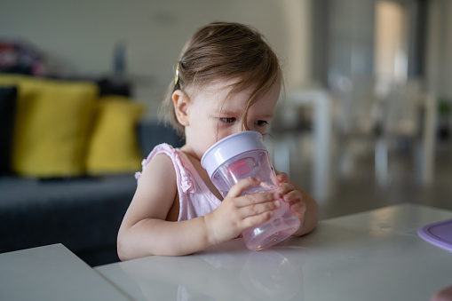 One child small caucasian toddler female baby holding plastic cup drink water alone at the table at home in room real people copy space early child development concept learning side view
