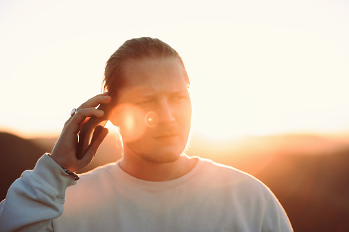 Bad News on Mobile Phone. Young blonde man in white shirt standing outdoors having a phone call with his Girlfriend on mobile phone, looking skeptical and can`t belief the news he just heard. Backlit from the sunset sun. Real People Millennial Generation Outdoors Lifestyle Portrait.