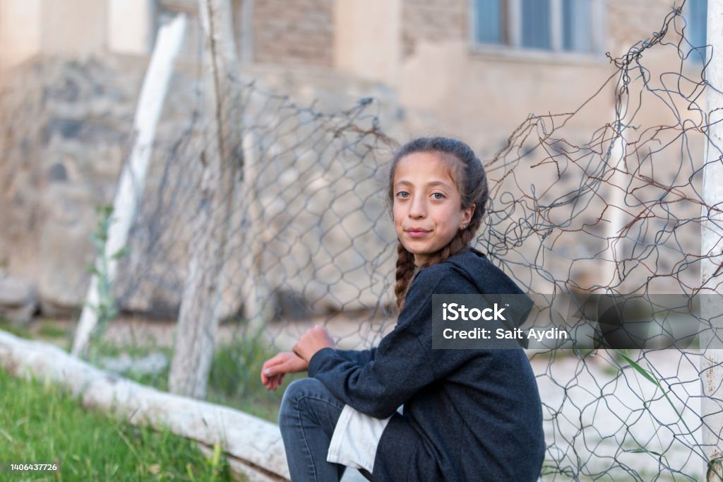 Refugee girl sitting near wire mesh fence. There is a blond blue-eyed girl in the photo. The girl is wearing black clothes. There are wire mesh fences behind the girl. Refugee Stock Photo