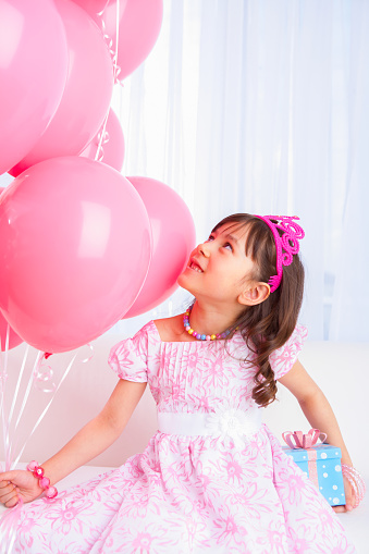 Smiling 8 years girl all dress up celebrating her birthday holding a group of pink balloons sitting on white sofa in living room.
