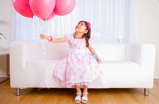 Smiling 8 years girl all dress up celebrating her birthday holding a group of pink balloons sitting on white sofa in livingroom.