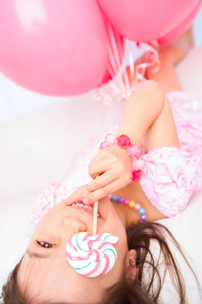 Smiling 8 years girl all dress up celebrating her birthday eating lollipop laying on white couch.