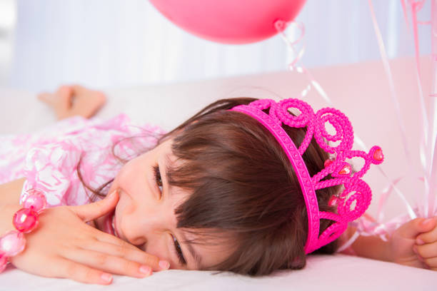 Smiling 8 years girl all dress up celebrating her birthday holding a group of pink balloons laying on white sofa in living room.
