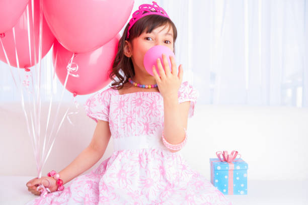 8 years girl all dress up celebrating her birthday blowing balloon sitting on white sofa in living room.