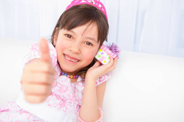 Smiling 8 years girl all dress up celebrating her birthday gesturing the thumbs up sitting on couch.
