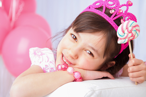 Smiling 8 years girl all dress up celebrating her birthday holding her lollipop laying on white couch.