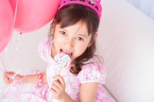 Smiling 8 years girl all dress up celebrating her birthday eating lollipop sitting on white couch.