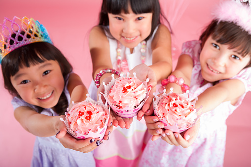Smiling three girls best friends celebrating birthday on pink background offering cupcakes.