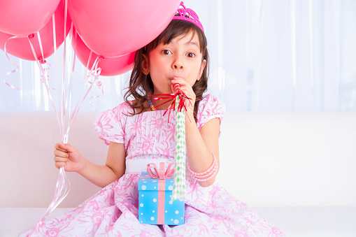 8 years girl all dress up celebrating her birthday blowing balloon sitting on white sofa in living room.