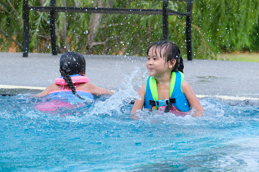 Asian child playing in the pool. Wearing orange life jacket, smiling with thumbs up.
