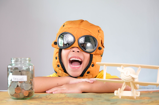 Smiling little girl in a pilot hat with coins in a clear jar for savings for travel and a wooden toy plane on the table. Childhood dream imagination and Travel concepts.
