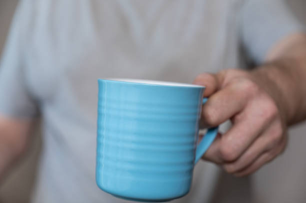 istock A middle-aged man is holding a blue mug. A ceramic cup with a handle for hot drinks in a man's hand. Middle section of a mature man in a gray T-shirt. Inside the room. Selective Focus. 1406452651