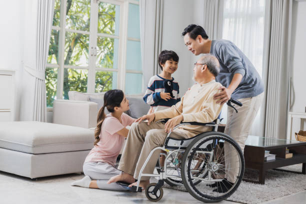 Happy family of senior with beautiful daughter, her husband and grandson take care old man sitting on wheelchair in house, positive dad have strength and positive thinking stock photo