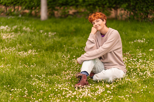 Positive androgynous female in casual outfit leaning on hand and looking at camera, while resting on grassy meadow with white flowers on summer day in park