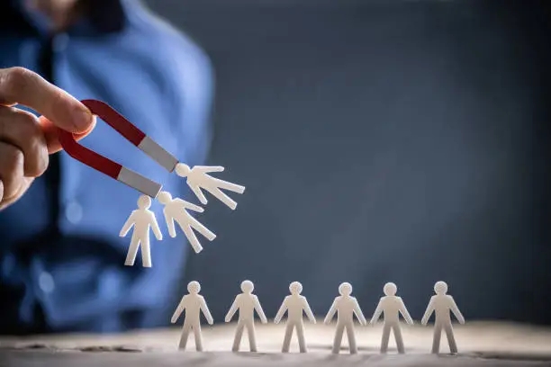 Close-up Of A Human Hand Attracting Red Human Figures With Horseshoe Magnet On White Background