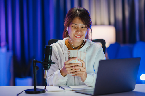 Young Asian woman holding money bank note and recording a podcast on her laptop in home studio at night. Podcaster concept.