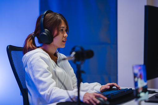 Young Asian woman playing online computer video game and broadcast streaming live in neon lights living room at home. Gamer lifestyle concept.