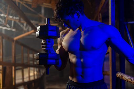 Strong Asian sportsman do sport workout weight training exercise in abandoned building. Shirtless man bodybuilder athlete practicing muscular strength bodybuilding lifting heavy dumbbell in dark old gym