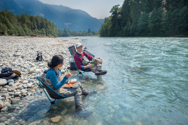 60+ Senior Asian Couple Eating Picnic Lunch While River Fishing stock photo