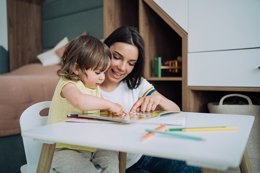 Shot of a little girl sitting at a table and coloring in a coloring book while her mother helping her. Mother teaching her daughter drawing and painting. Mom spending time with her daughter. Happy family at home.