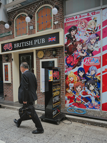 Tokyo, Japan - 02 02 2019. Elderly man passes in front of a fake Japanese British Pub next to a billboard with manga characters.