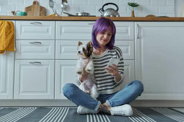 Young woman spending time with dog while using phone and sitting on the floor at the kitchen stock photo