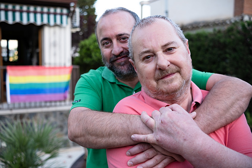 Bearded middle aged man embracing husband from behind and looking at camera while spending time in yard of house decorated with LGBT flag