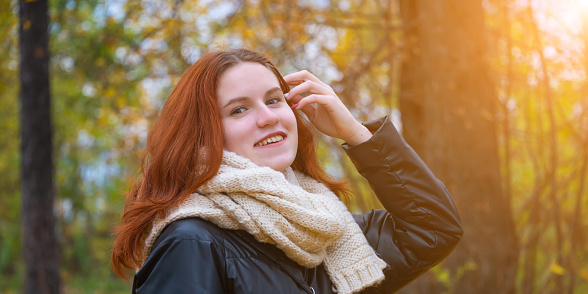 portrait of a red-haired smiling girl in a jacket and scarf straightening her hair. against the background of autumn nature and the bright sun, the concept of human emotion.