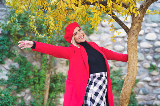 Cheerful young elegant woman with blond hair and red lips in stylish coat and beret holding tree trunk, while standing in autumn park with outstretched arm and smiling