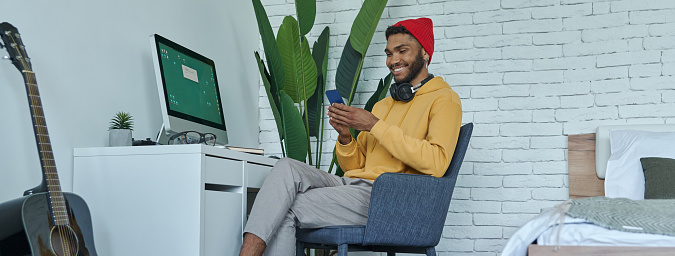 Happy young African man using smart phone while sitting at his working place at home